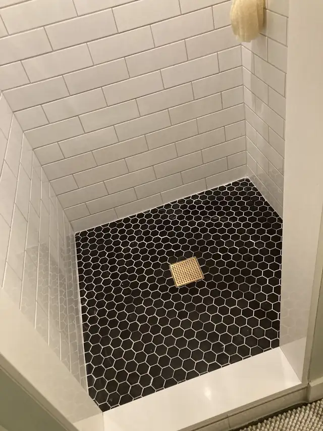 Shower floor in black hexagon mosaic tile with a square brass drain and white subway tile walls