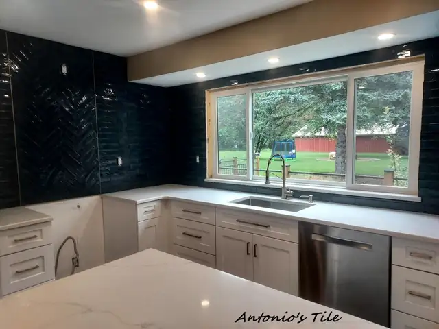 Kitchen with navy herringbone tile backsplash, white shaker cabinets and white countertop under a wide window